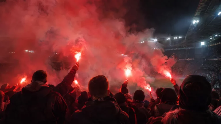 Fußballfans in einer Stadionkurve zünden nachts rote Bengalos, dichter Rauch zieht über die Tribüne.