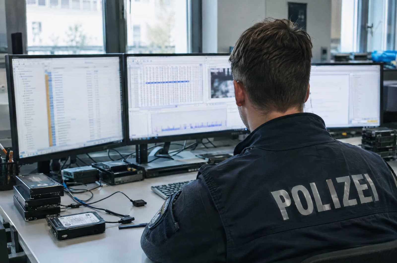 Ermittler der IT-Forensik sitzt an mehreren Monitoren und wertet sichergestellte Datenträger aus einem Chatgruppen-Verfahren aus.