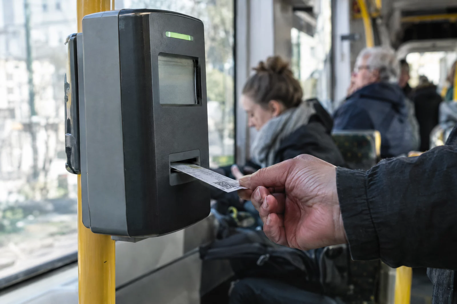 Eine Hand hält einen Papierfahrschein an den Entwerter in einer Straßenbahn, im Hintergrund unscharf andere Fahrgäste.