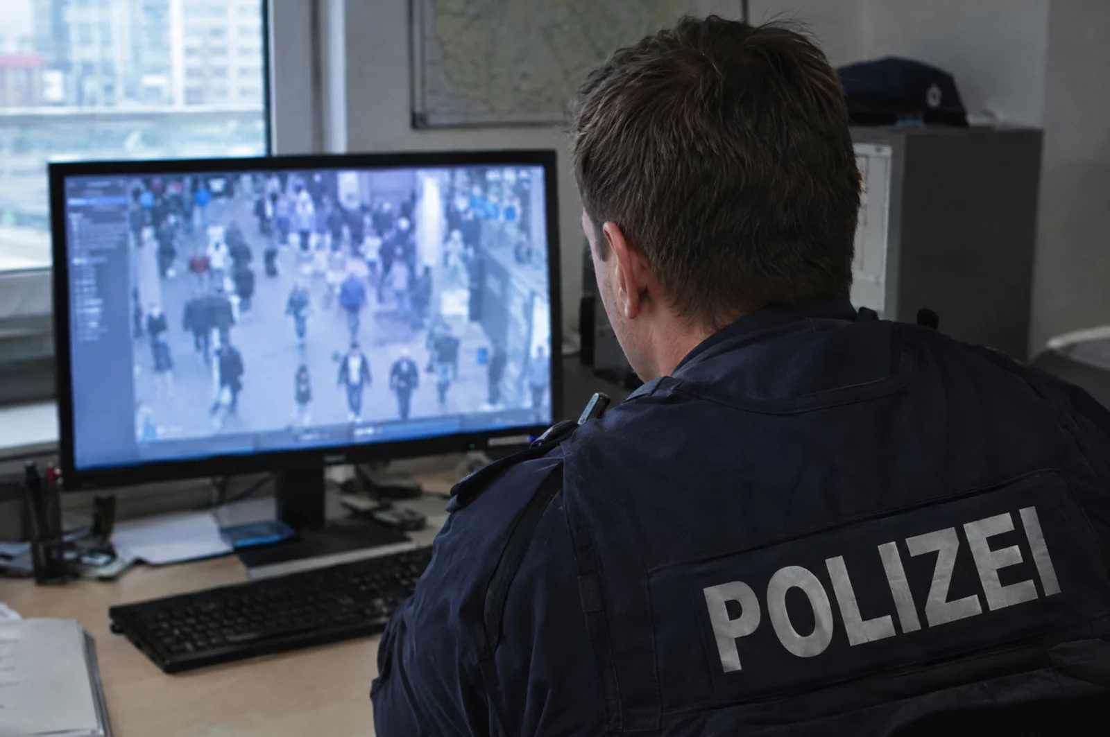 Polizeibeamter sichtet am Monitor unscharfe Überwachungsaufnahmen einer Menschenmenge, etwa zur Klärung einer Schlägerei.