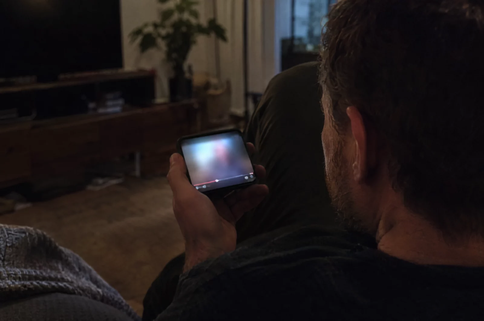 Mann schaut im dunklen Wohnzimmer auf ein Smartphone mit unscharfem Videoplayer, Thema Verbreitung von Gewaltdarstellungen.