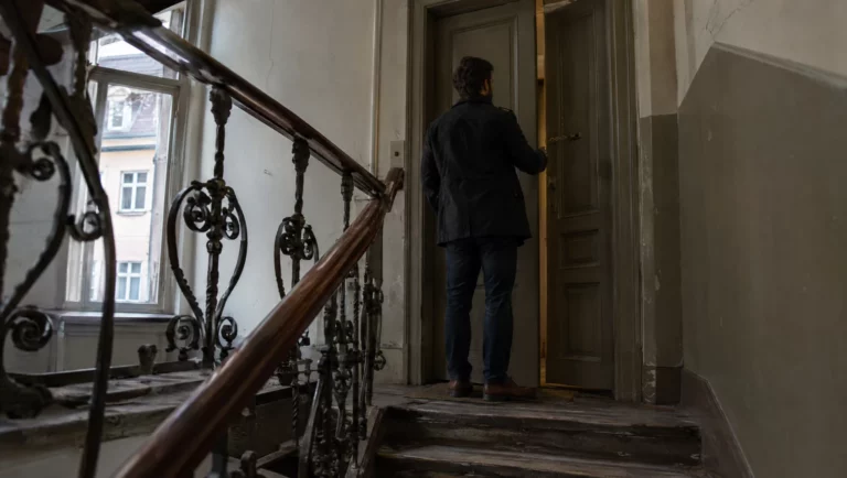Ein Mann steht im Altbau-Treppenhaus vor einer Wohnungstür, die nur mit vorgelegter Kette geöffnet ist – Thema Hausfriedensbruch.