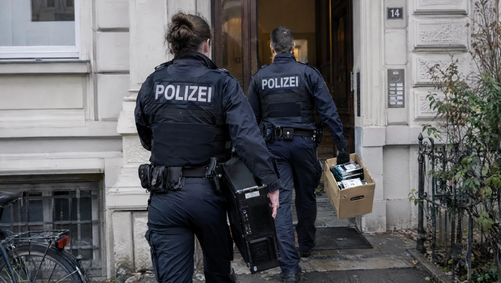 Zwei Polizeibeamte tragen einen Computer und einen Karton mit Datenträgern am Wohnhaus, Durchsuchung wegen KI-generierter Inhalte nach § 184b StGB.