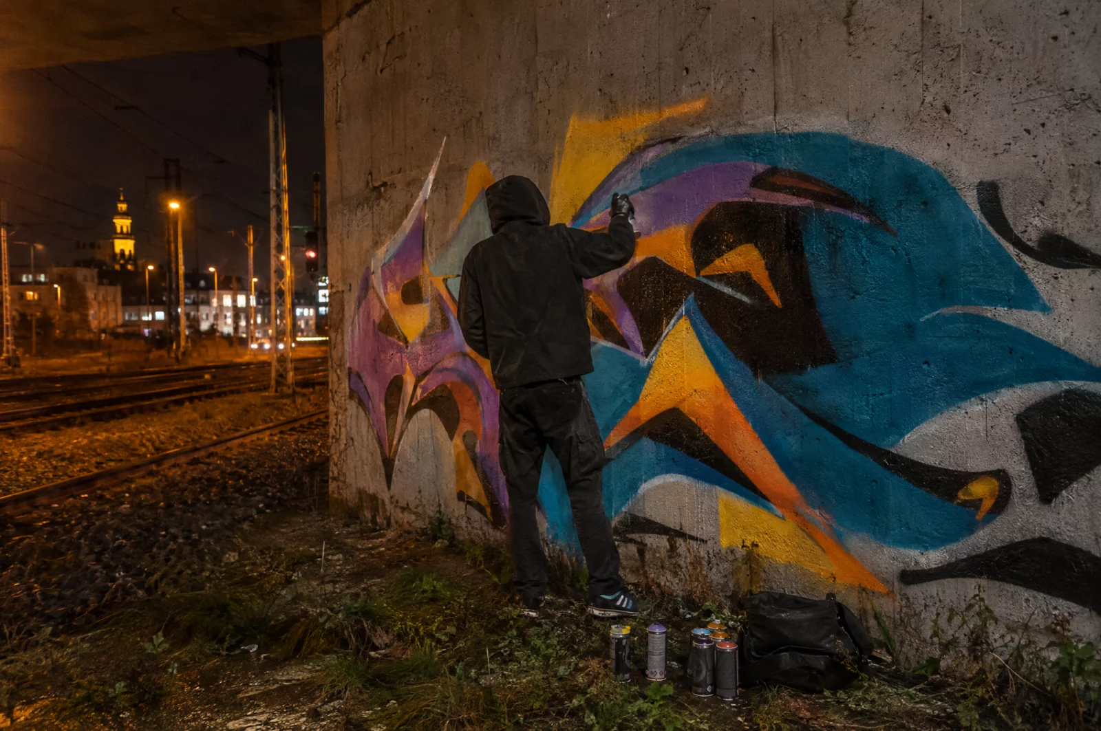 Eine Person sprüht nachts ein buntes Graffiti an die Wand einer Bahnunterführung, Spraydosen stehen am Boden.