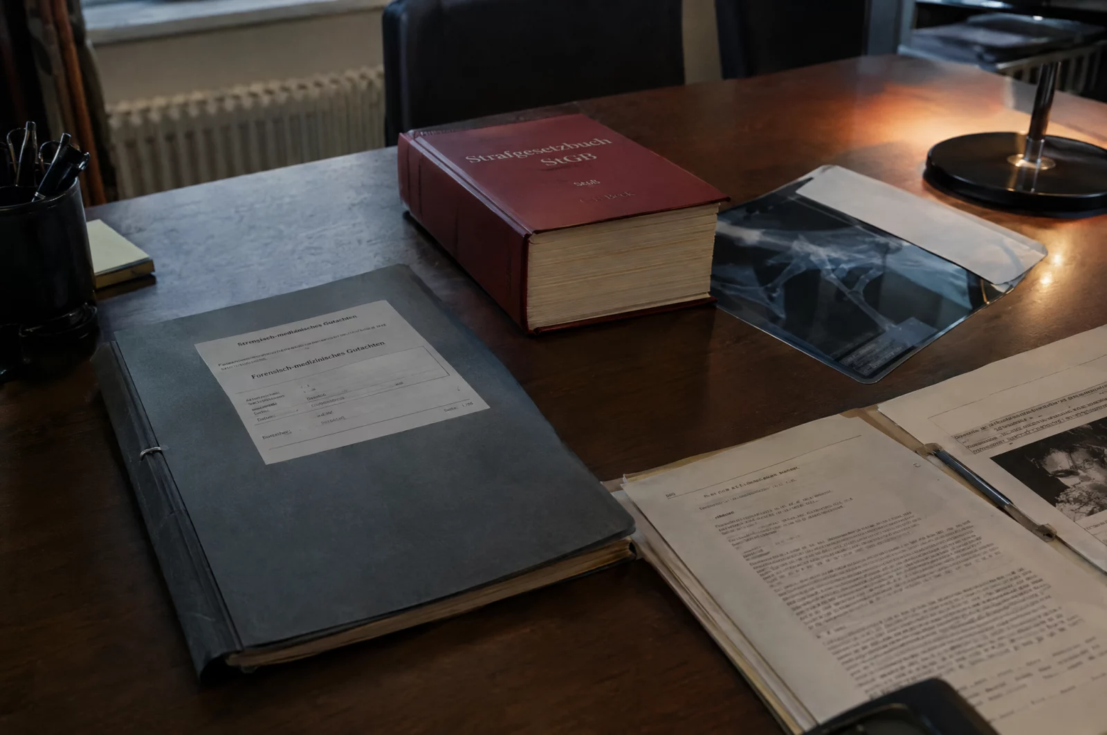 Rechtsmedizinisches Gutachten in grauer Mappe neben Strafgesetzbuch und Röntgenumschlag auf dem Schreibtisch eines Verteidigers.