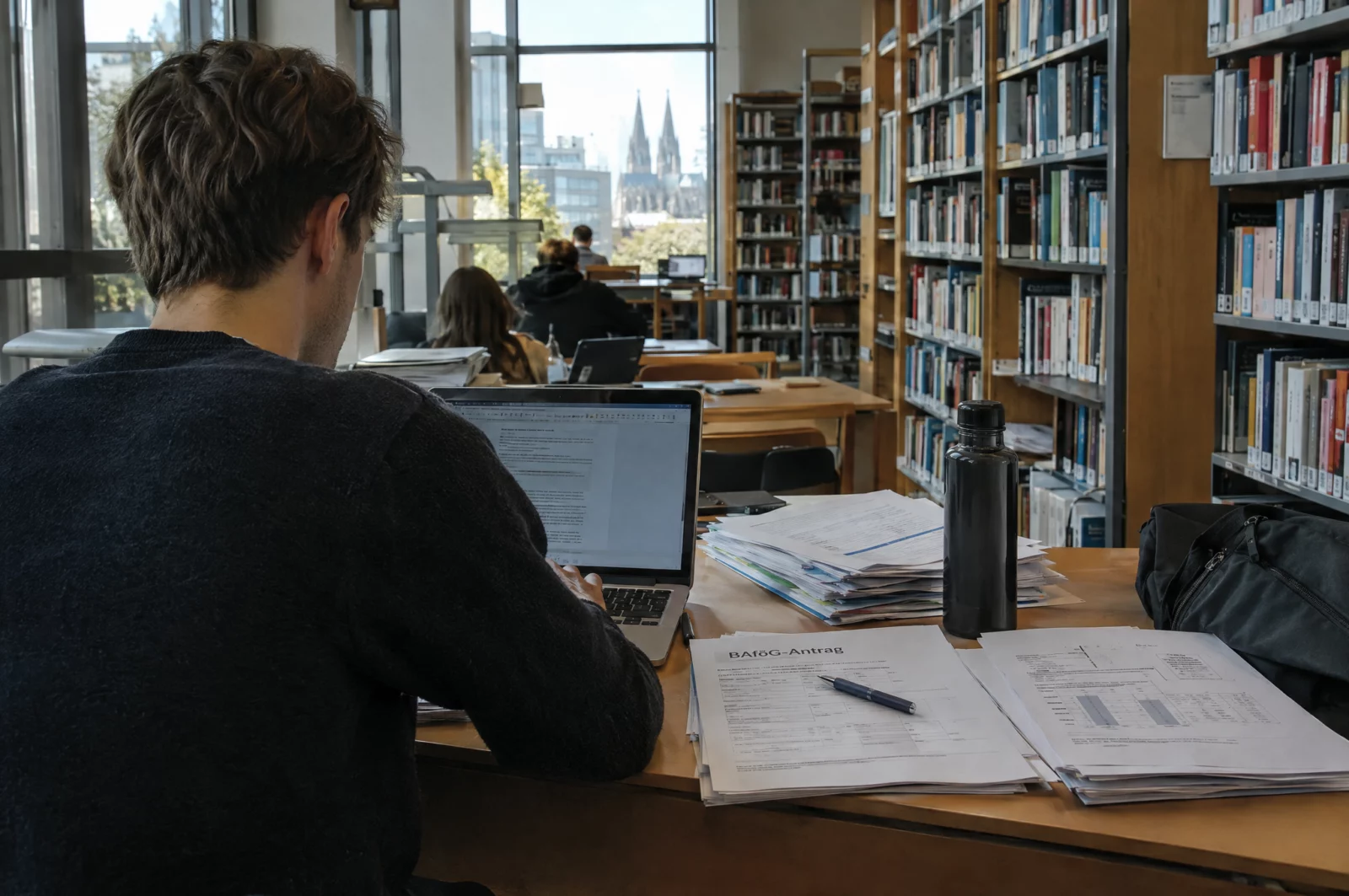 Sozialbetrug - § 263 StGB Student sitzt in einer Universitätsbibliothek mit Laptop und Finanzunterlagen für seinen BAföG-Antrag.
