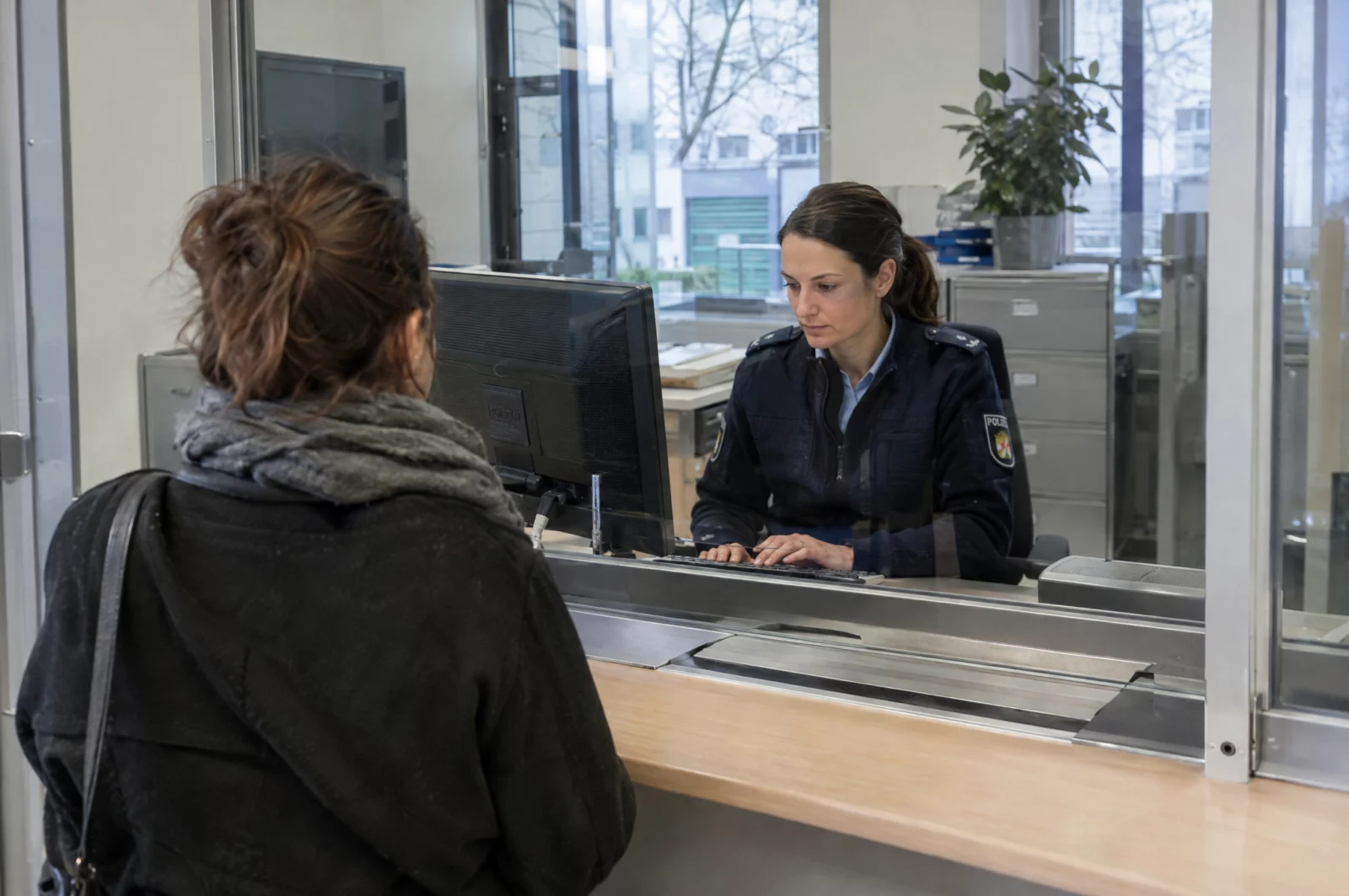 Polizeibeamter nimmt hinter einer Glasscheibe auf der Wache eine Strafanzeige auf, der erste Schritt im Gang des Strafverfahrens.